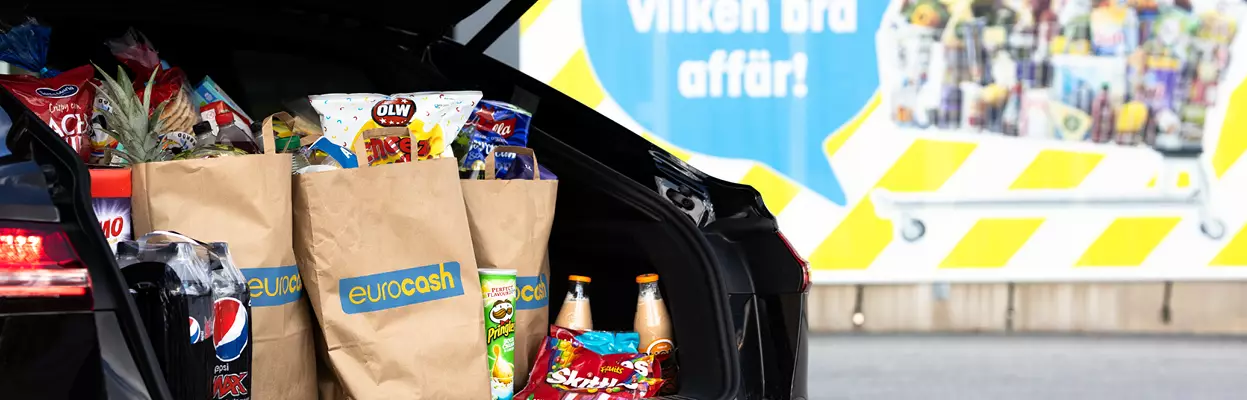 Fullpackade matkassar från eurocash med mat placerade i backluckan på en bil. I bakgrunden hänger en fasadvepa med bild på Eurocash kundvagn och budskapet "WOW vilken bra affär".