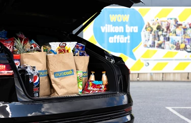 Fullpackade Eurocash papp-påsar med mat i bakluckan på en bil. I bakgrunden hänger en reklam-banner med budskapet "WOW vilken bra affär!"