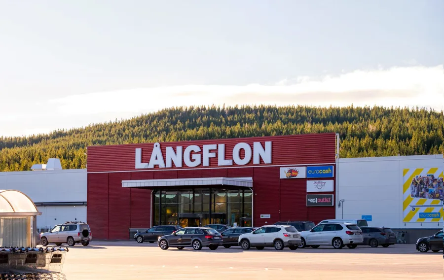 Långflon shoppingcenters fasad och parkering en solig dag. I bakgrunden står den vackra granskogen på ett fjäll.